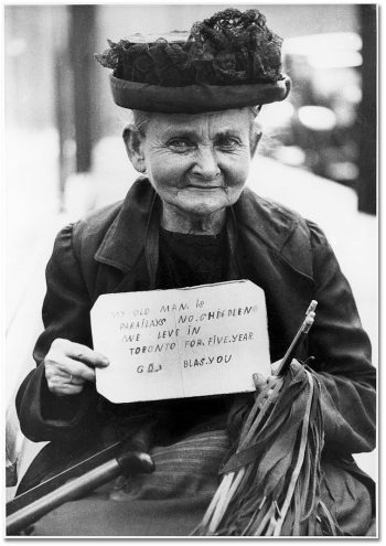 Elderly woman in black lace hat holds up misspelled sign that reads: My old man is paralyzed, no children, we live in Toronto for five year, God bless you.