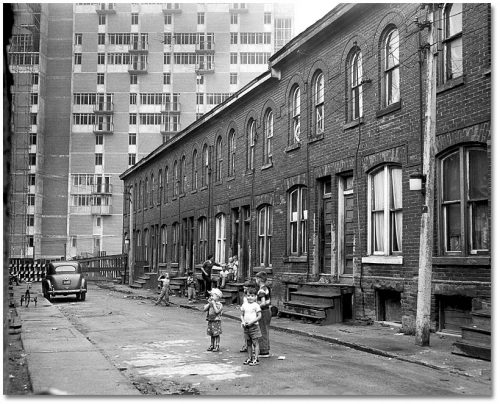 Children play on a narrow street lined with old brick houses, while a highrise takes up the background.