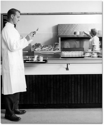 Standing in front of a cafeteria food line, a man in a white lab coat examines a spoon.