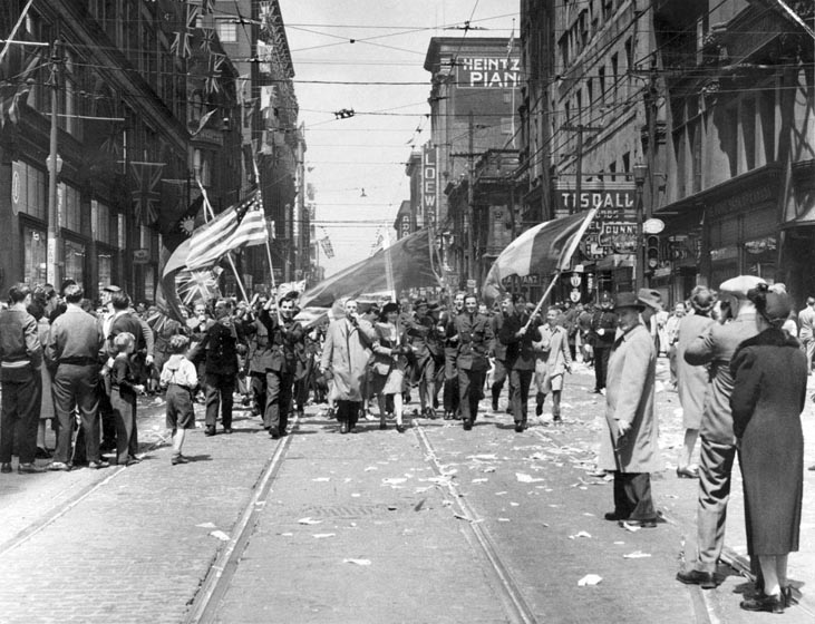 Armed forces personnel and civilians are parading down Yonge Street, waving the Union Jack and American and Italian flags.