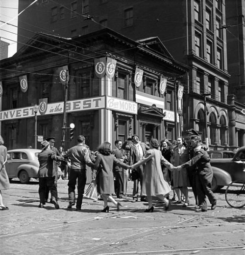 People are dancing in a circle in an intersection. The building behind them is covered in ads for Victory Bonds.