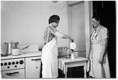 Beside a stove, a woman watches another pour milk from a pitcher into a bottle.