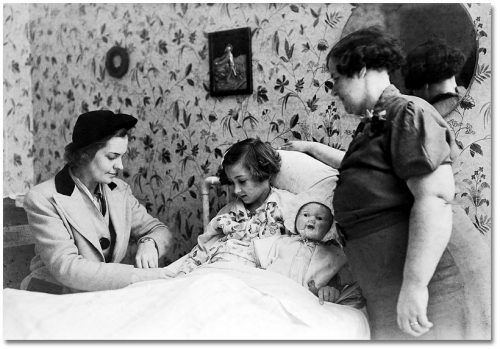 A woman takes the pulse of a girl who is sitting in bed with a large doll beside her.