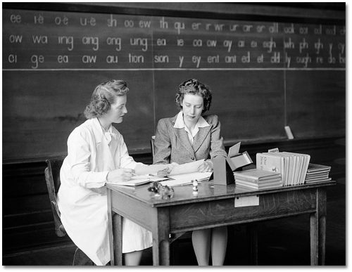 Two women are talking at a desk in front of a blackboard.