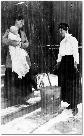 Two women, one holding a baby, stand outside looking at a garbage can.