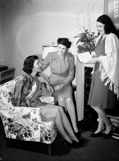 Three women are gathered in a living room. They are all wearing skirts, but one wears a jacket draped over her shoulders, the second wears a suit with jacket fastened in front, and a third has a shawl over her dress.