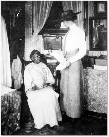 A white woman stands beside a Black woman sitting in a chair.