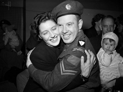 A soldier and a woman embrace cheek to cheek. A baby looks on from the side.