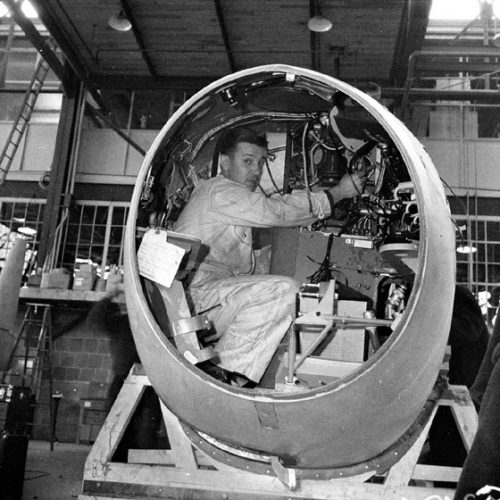 A man works on wiring while squatting in an airplane's oval cockpit.