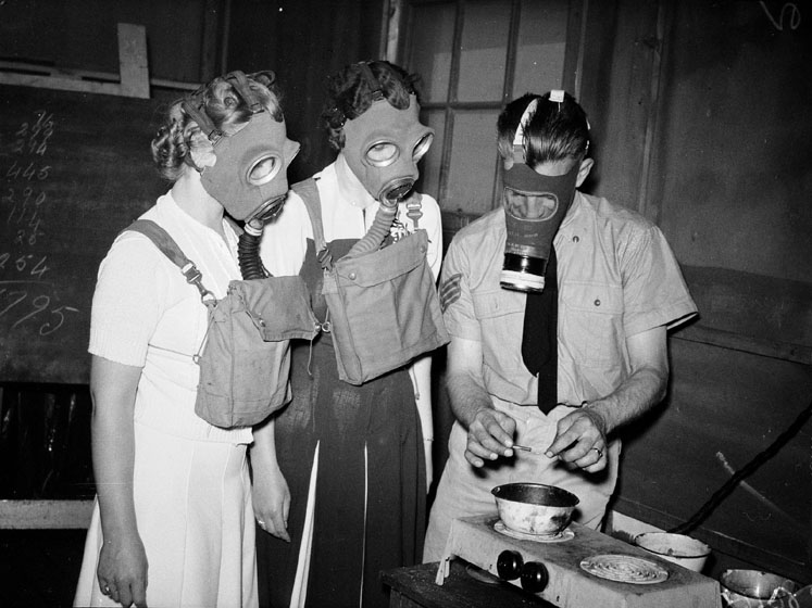 A soldier cooks something in a small bowl on a hotplate. Two women in civilian clothes watch him. All are wearing gas masks.