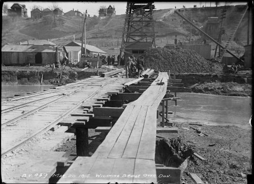 Temporary wooden bridge over river leading to excavation equipment and piles of earth.