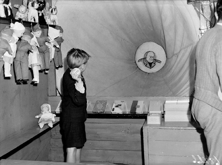 A boy throws a ball at a target showing a caricature of Mussolini. Prizes are lined up in front of the boy and also hanging on the wall at his side.