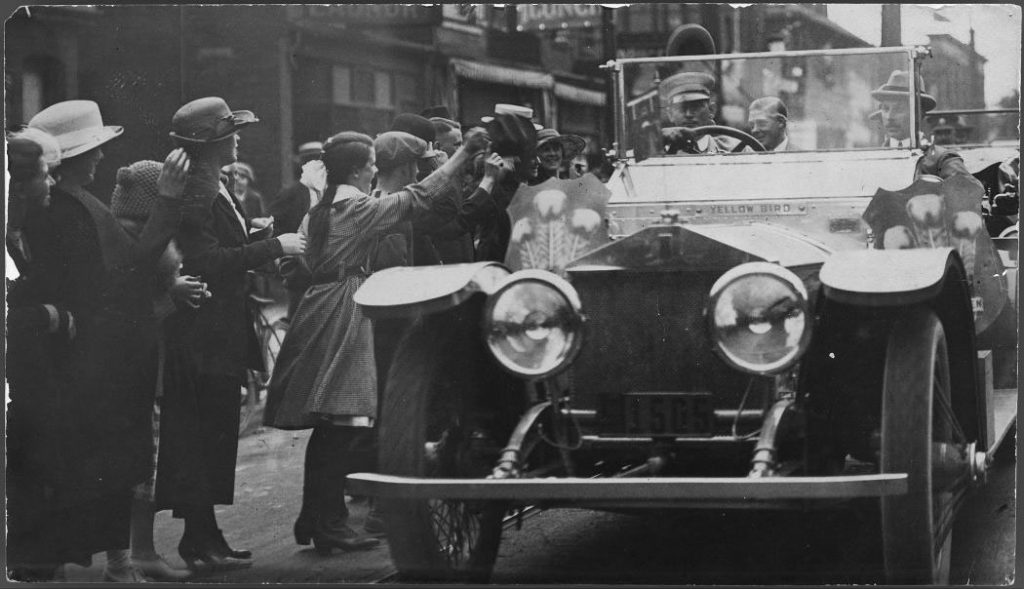 Girls at the side of the road waving to man in back of open-topped car.