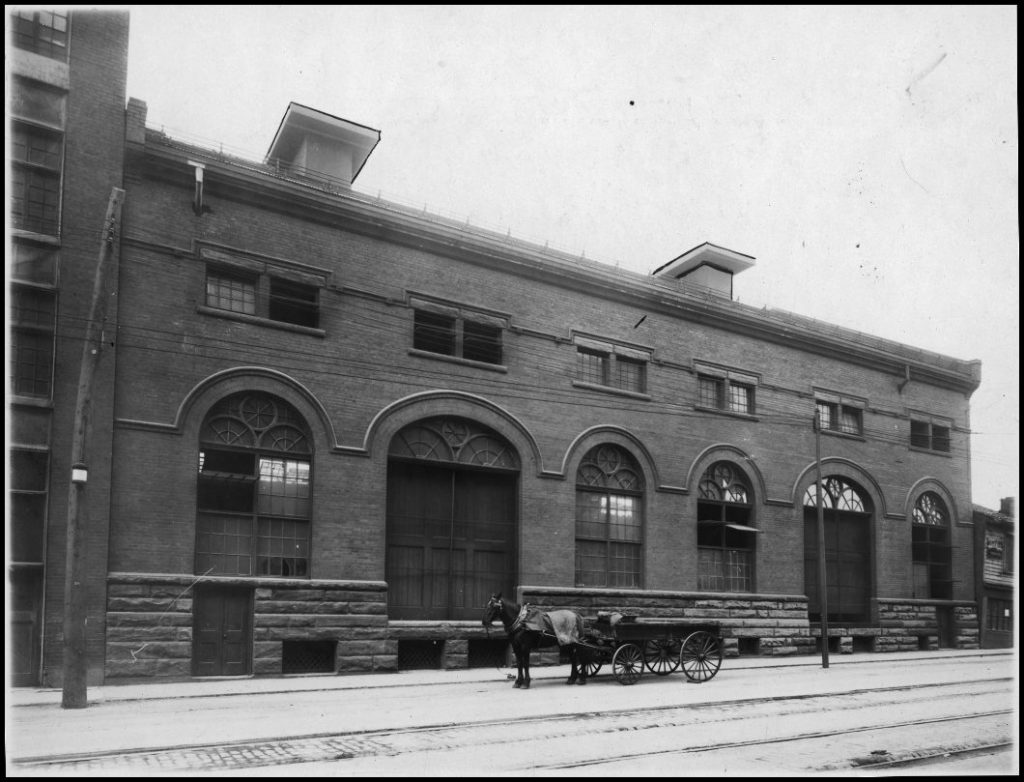 A brick building with wide arched doors, and a horse and cart waiting in front.