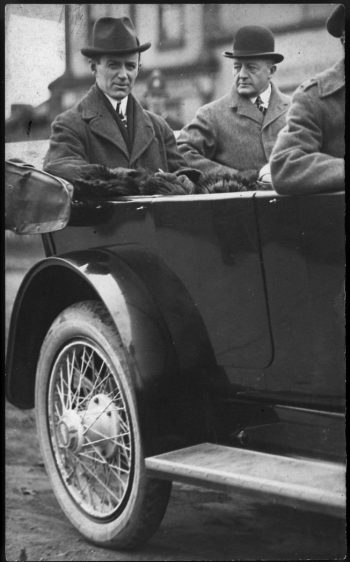 Two men in hats in the back of an open-topped car.