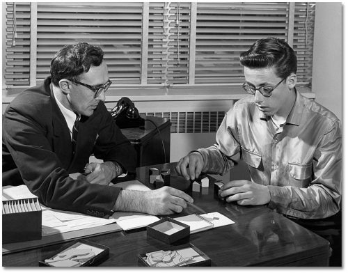 A man points at a diagram in a notebook while a younger man tries to recreate the diagram using wooden blocks.