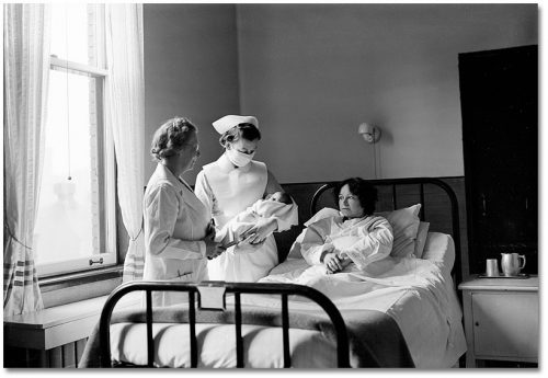 Woman lies in hospital bed while a nurse beside the bed holds a baby and another nurse looks on.