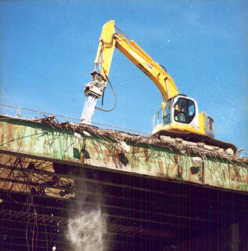 Large yellow machine hammers at top of old deck, and concrete dust falls below