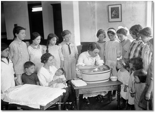 Young teenage girls watch a woman bathe a baby in a basin on a table.