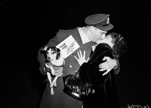 A soldier kisses a woman. In his arm he holds a small dog that has a card attached to it reading 'Welcome home George!'