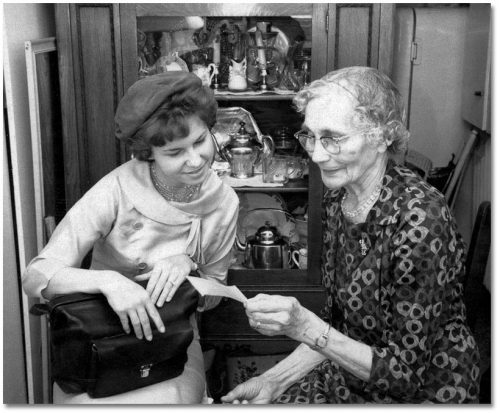 A young woman in a hat and an elderly woman in a patterned dress look together at a photograph.