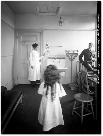 A girl stands at one end of a room, while at the other, a woman points to an eye examination chart.