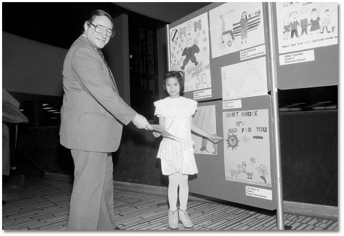A man shakes a girl's hand in front of a poster display. She is pointing to a poster with drawings of a sun and flowers. It says Don’t smoke it's bad for you.