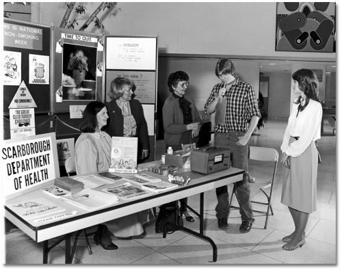 In front of a display of anti-smoking posters and brochures, a young man breathes into a device while people watch.