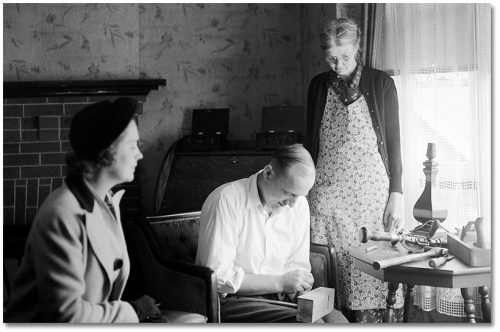 A nurse sits with an elderly woman wearing an apron, and an elderly man who seems to be doing some sort of woodworking with a block of wood in his lap.