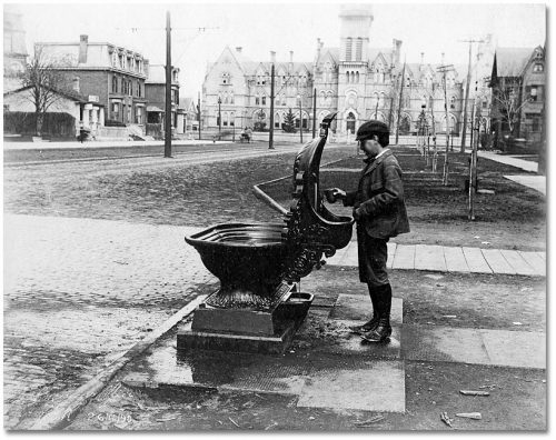 A boy fills a cup in front of a cast iron fountain that has a spout for humans, a large basin for horses, and a smaller basin for dogs.