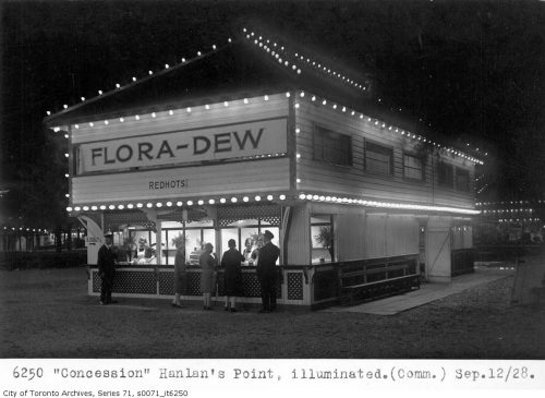 People line up at a two-storey wooden concession building that is outlined on all corners with lights.