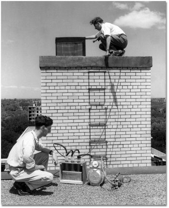 Two men on a roof use a machine with a motor and coils.