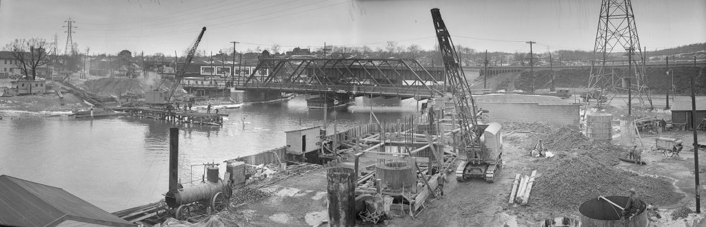 Construction on a pier in a river, with construction equipment and concrete forms, and a steel bridge in the background.