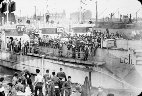 Several troop carriers are moored side by side. Soldiers and sailors crowd the decks, standing and sitting, talking and smoking.