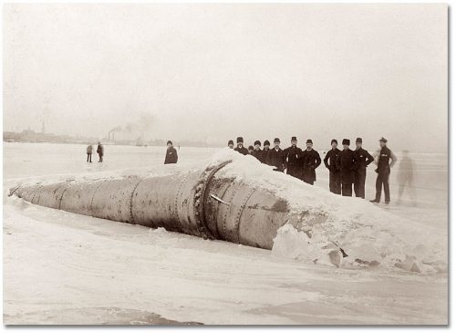 Large pipe rests on ice on the bay, broken in half, with men standing around looking at it.
