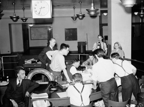 Reporters stand aroud a semicircular desk littered with papers. Threre are also two phones on the desk. The clock hanging from the ceiling reads 2:34.