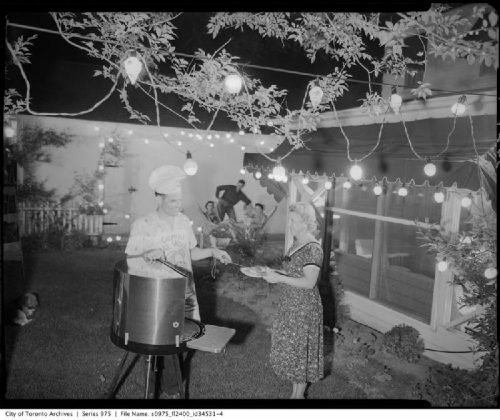 In a backyard illuminated with strings of lights, a man at a barbeque puts food on a plate held by a woman.