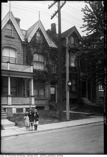 Three small girls stand on the sidewalk in front of a house. A streetlight is attached to the concrete pole beside them.