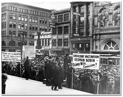 People on a platform speak to the crowd, who hold signs that say Stop the slaughter of the innocents, protest against compulsory vaccination and Compulsory vaccination is German born, down with compulsion.