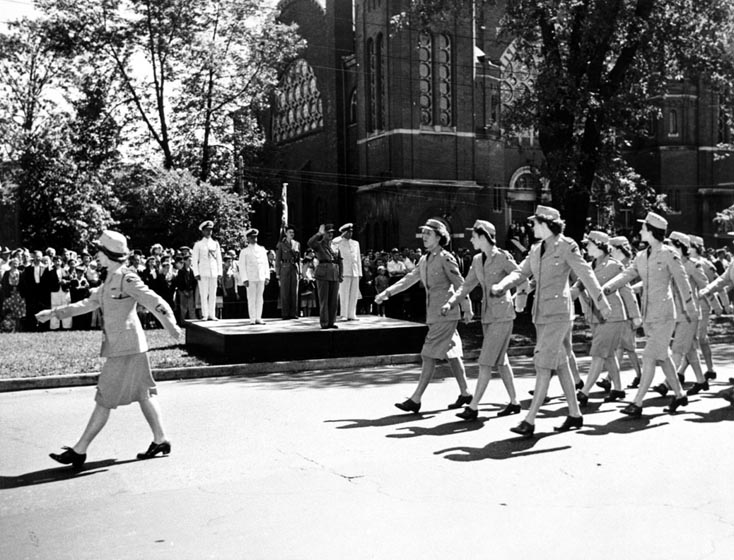 Women in uniforms march in formation past saluting officers.