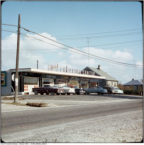 Cars parked in front of a strip mall with letters on the roof that say Evan’s Shopping Centre.