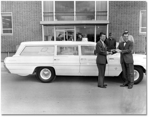 Two men in suit shake hands in front of a white station wagon-style vehicle.