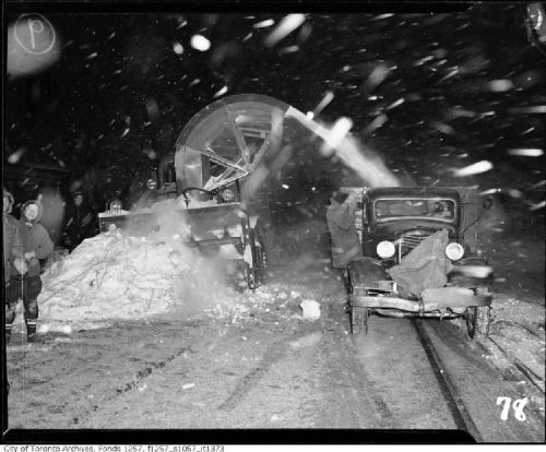 A snow blower vehicle shoots snow into the pack of a pickup truck. Smiling workers with shovels stand beside it.