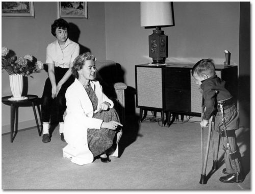 Small boy using leg braces and crutches walks towards two women in a living room.