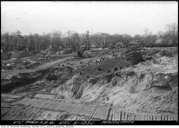 Machines clearing trees from section of land, with houses in the background.