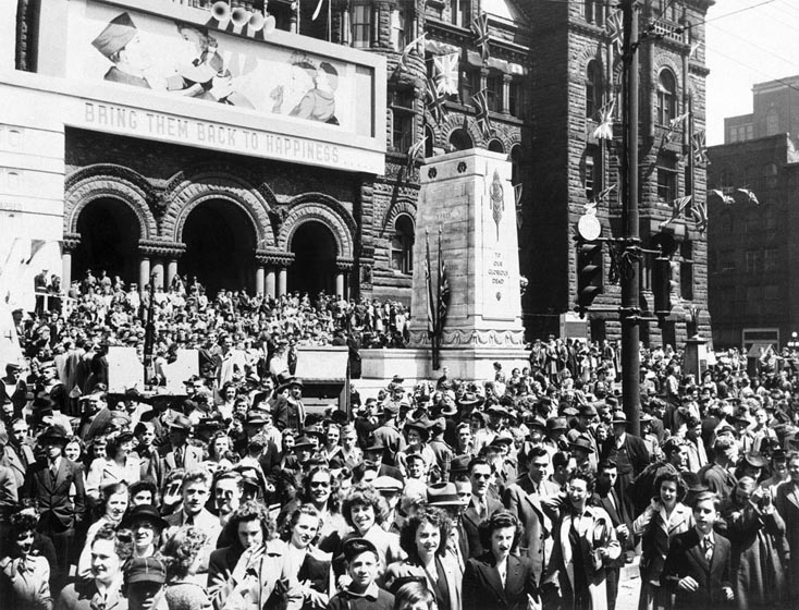 A crowd covers the area in front of Old City Hall. A sign on the City Hall shows a soldier and his family, and the words Bring Them Back to Happiness.