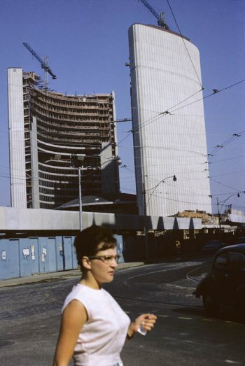 Woman walks in front of two curving towers of New City Hall under construction.