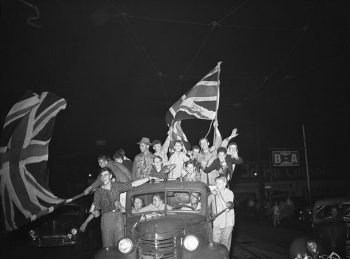 People crowded into the back of a pickup truck cheer and wave large Union Jacks.