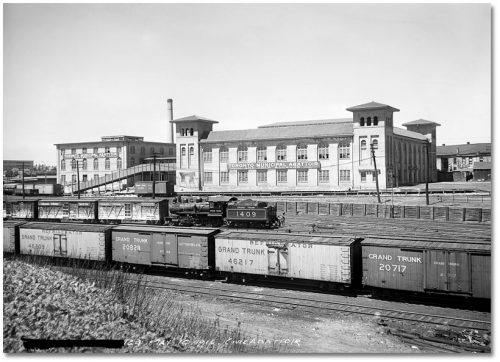 Two two-storey white brick buildings with large windows, and trains on railway tracks in front of them.