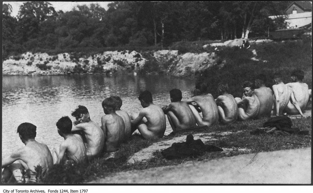 A group of naked boys with their backs to the camera sit on the bank of a river